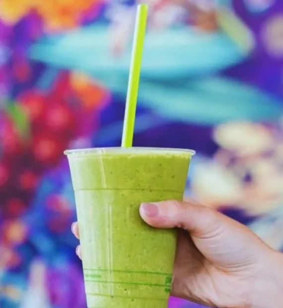 fresh green smoothie in a clear plastic cup with a lime green straw, set against a vibrant colorful floral background