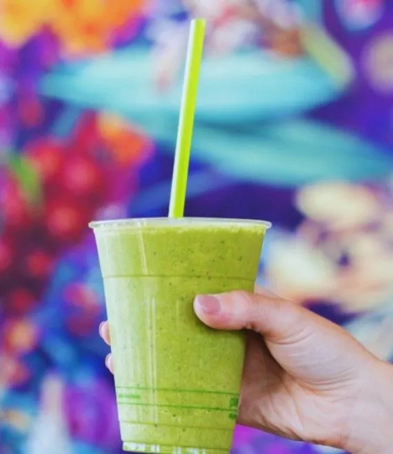 A hand holding a fresh green smoothie in a clear plastic cup with a lime green straw, set against a vibrant colorful floral background