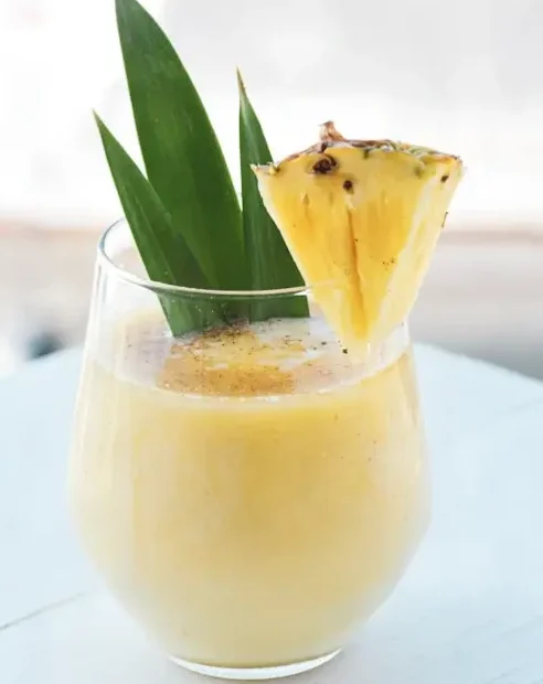 Fresh pineapple smoothie served in a glass with a pineapple wedge and tropical green leaves garnish on a white table.