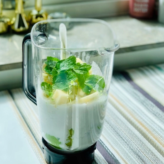 Close-up of a blender jar from filled with milk, pineapple chunks, and a large layer of fresh green mint leaves.