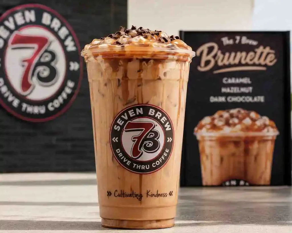 7 Brew Brunette iced coffee with caramel, hazelnut, and dark chocolate flavors topped with whipped cream in a branded clear cup.