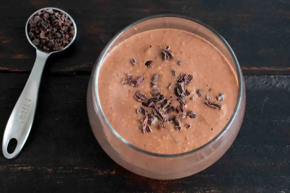 Top view of a creamy chocolate black bean smoothie topped with cacao nibs, served in a glass next to a tablespoon of cacao nibs on a wooden table.